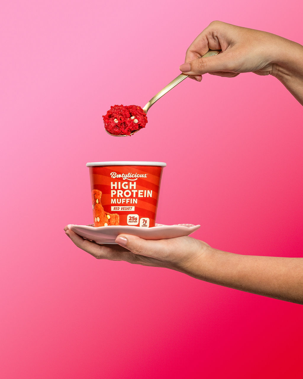 Hands holding a red velvet protein muffin cup on a pink backdrop